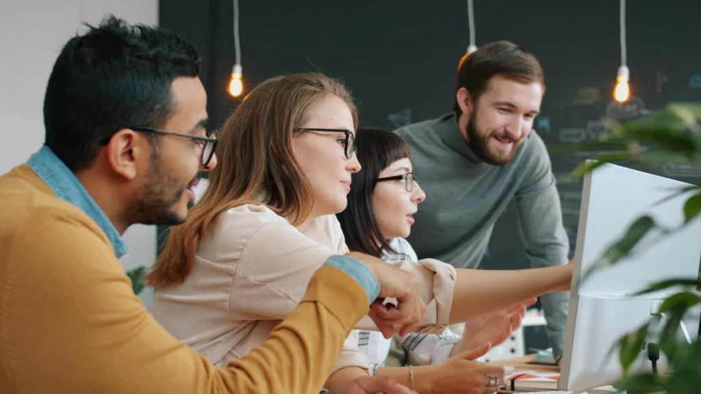 A diverse team of young professionals collaborating on a computer project in a modern office setting.